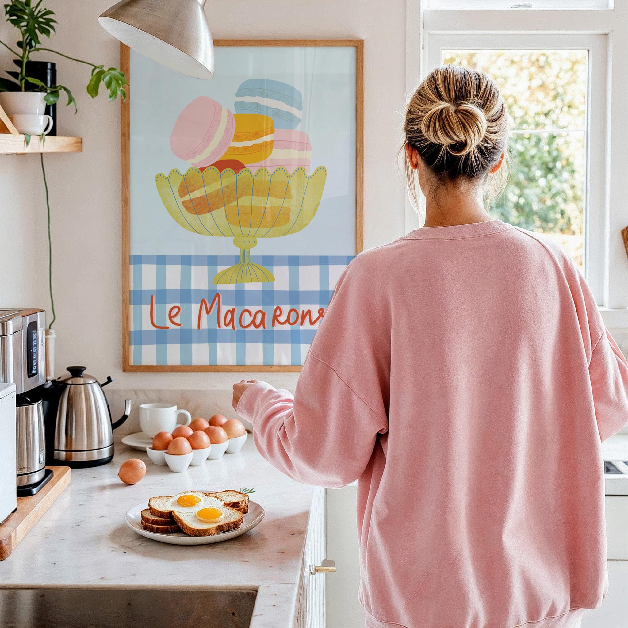 Person in a pink sweatshirt standing in a kitchen with a 'Le Macarons' poster on the wall.