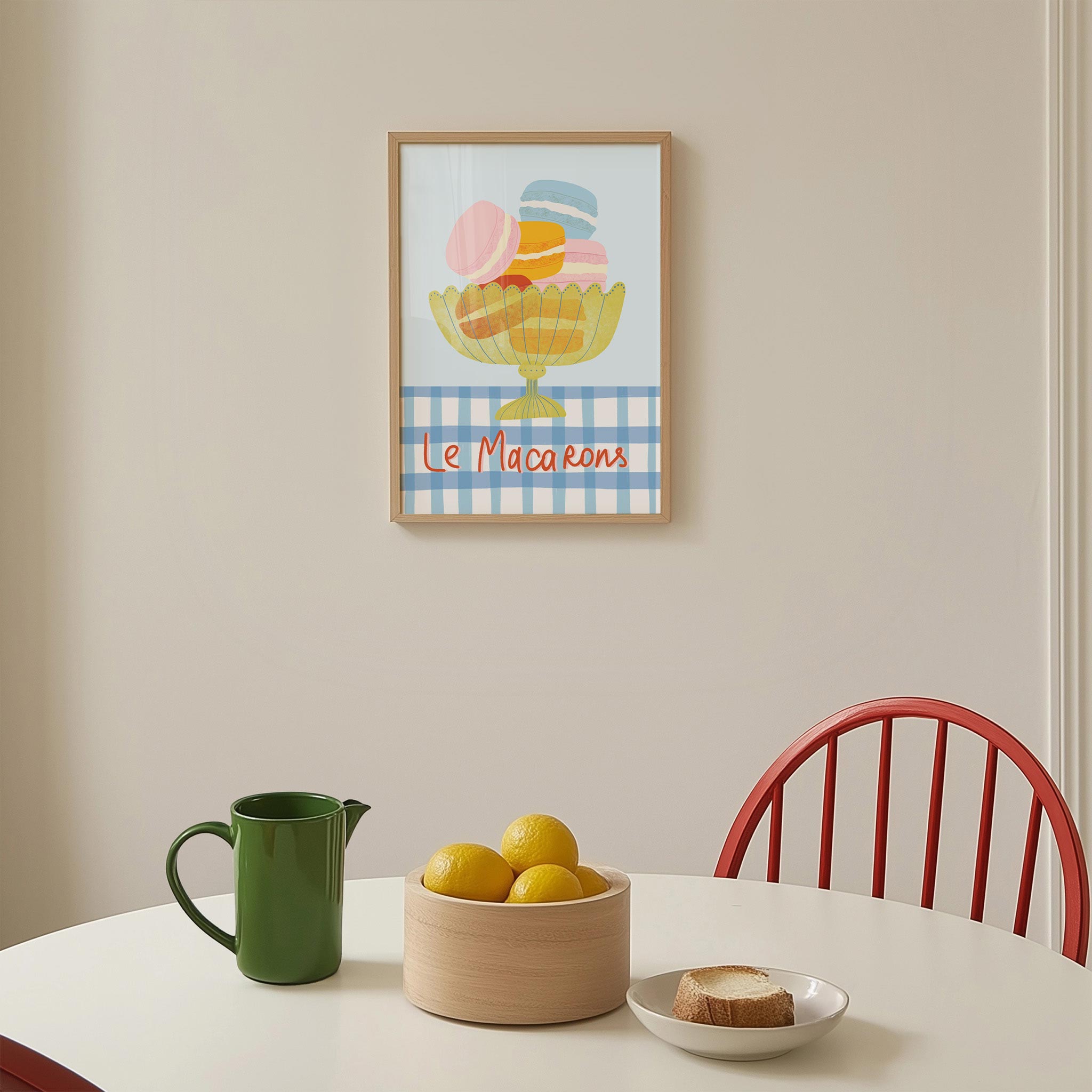 Framed artwork of macarons above a table with a pitcher, bowl of lemons, and plate of bread.